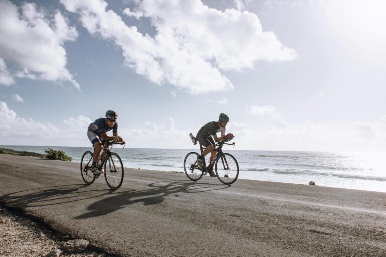 Two cyclists racing on a scenic coastal road under a bright, clear sky.